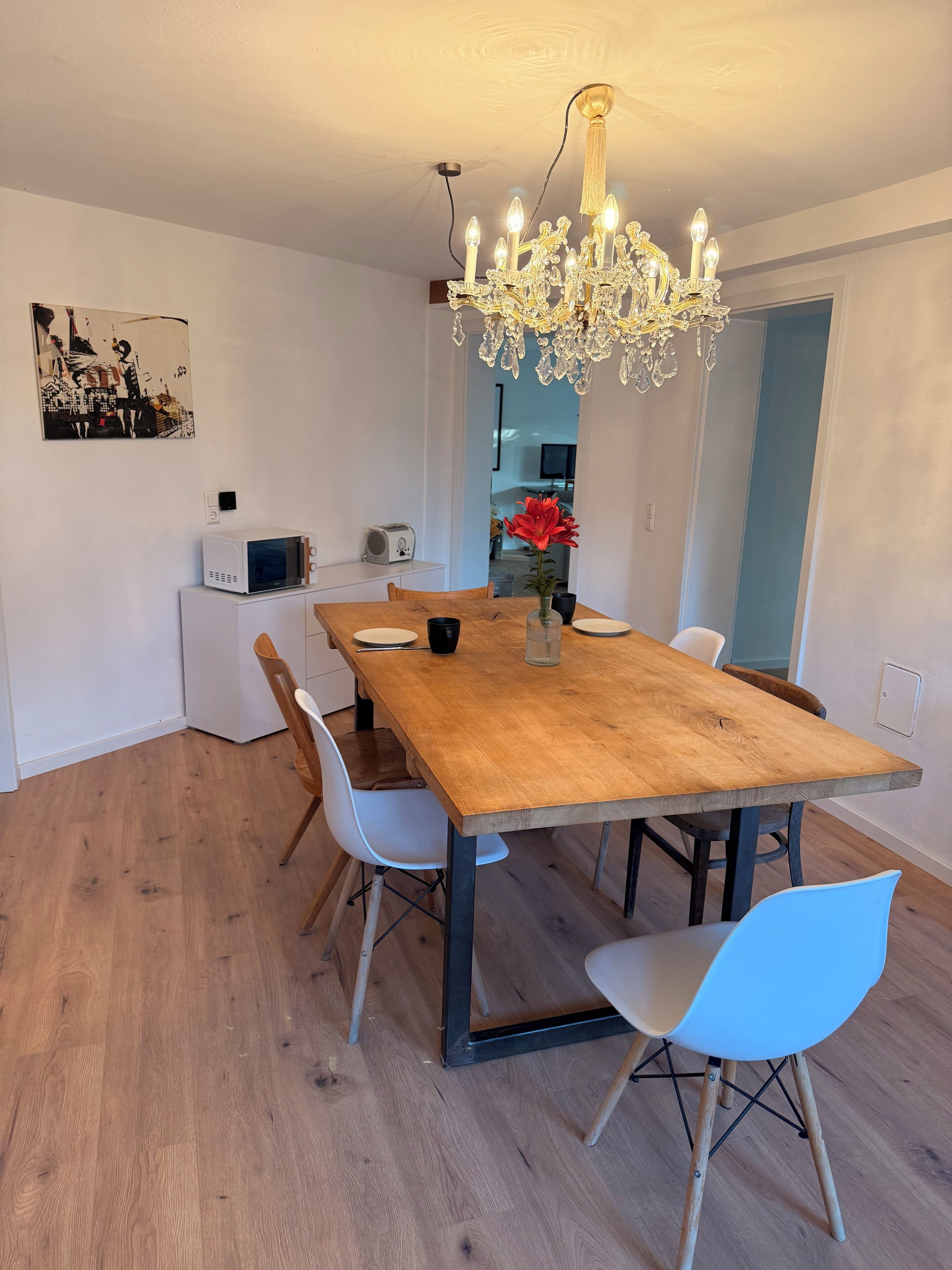 A dining area with a wooden table, white chairs, a chandelier, and a microwave on a cabinet.
