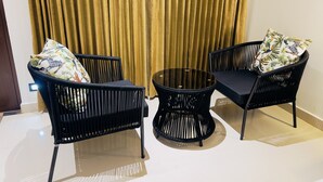 A room with two black wicker chairs with cushions, a small glass table, and a golden curtain in the background.