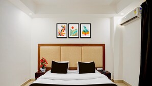 A hotel room with a bed, two bedside tables, a nightstand, a wall-mounted air conditioner, and three framed artworks above the bed.