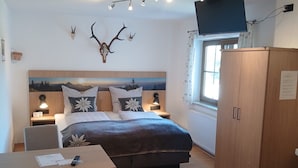 Ein Schlafzimmer mit Bett, Nachttischlampen, Schreibtisch, Stuhl, Kleiderschrank, einem aufgehängten Hirschkopf, einem Fernseher und einem Fenster.