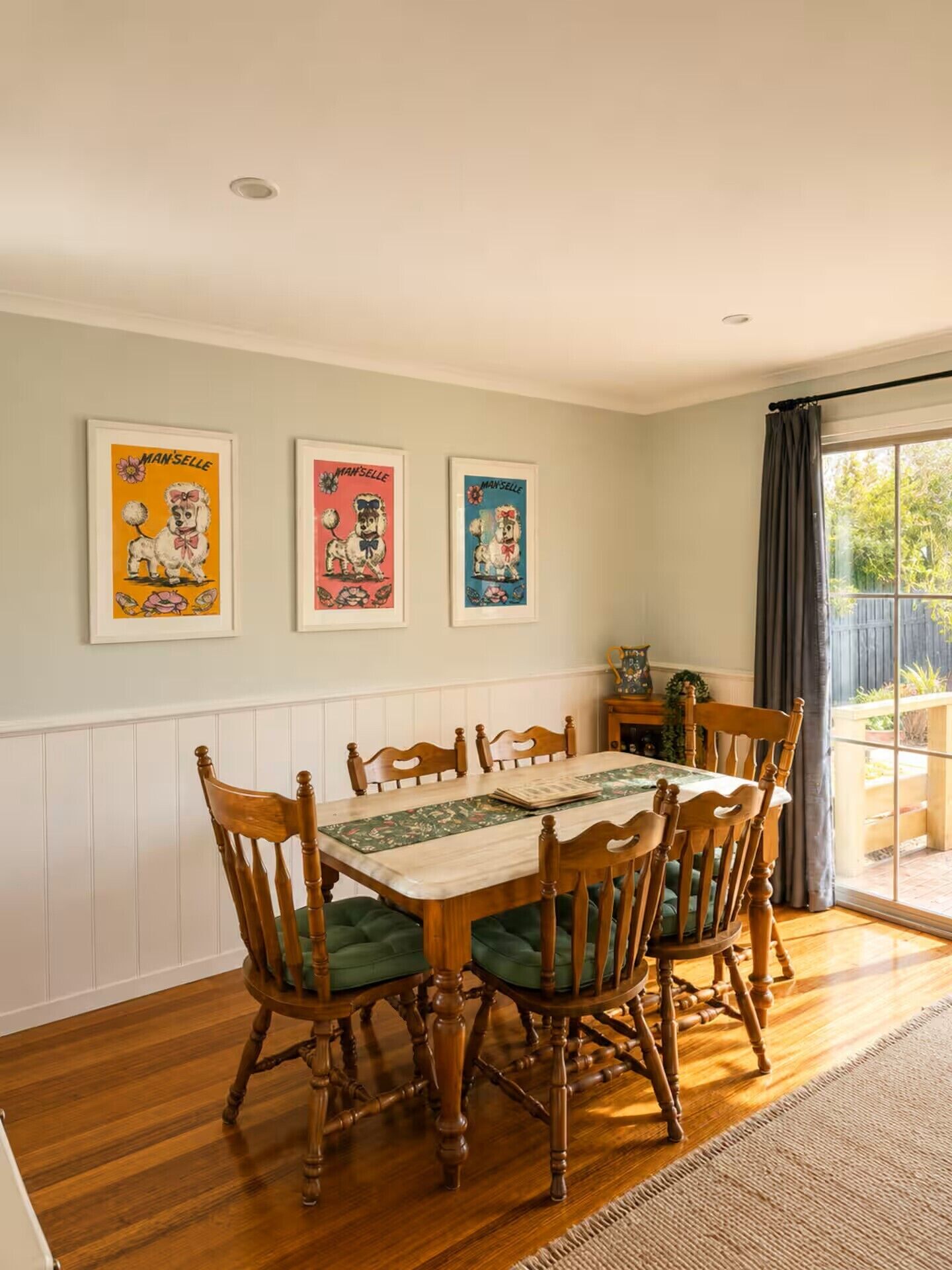 A dining room with a wooden table and chairs, framed posters on the wall, and a view through a window.
