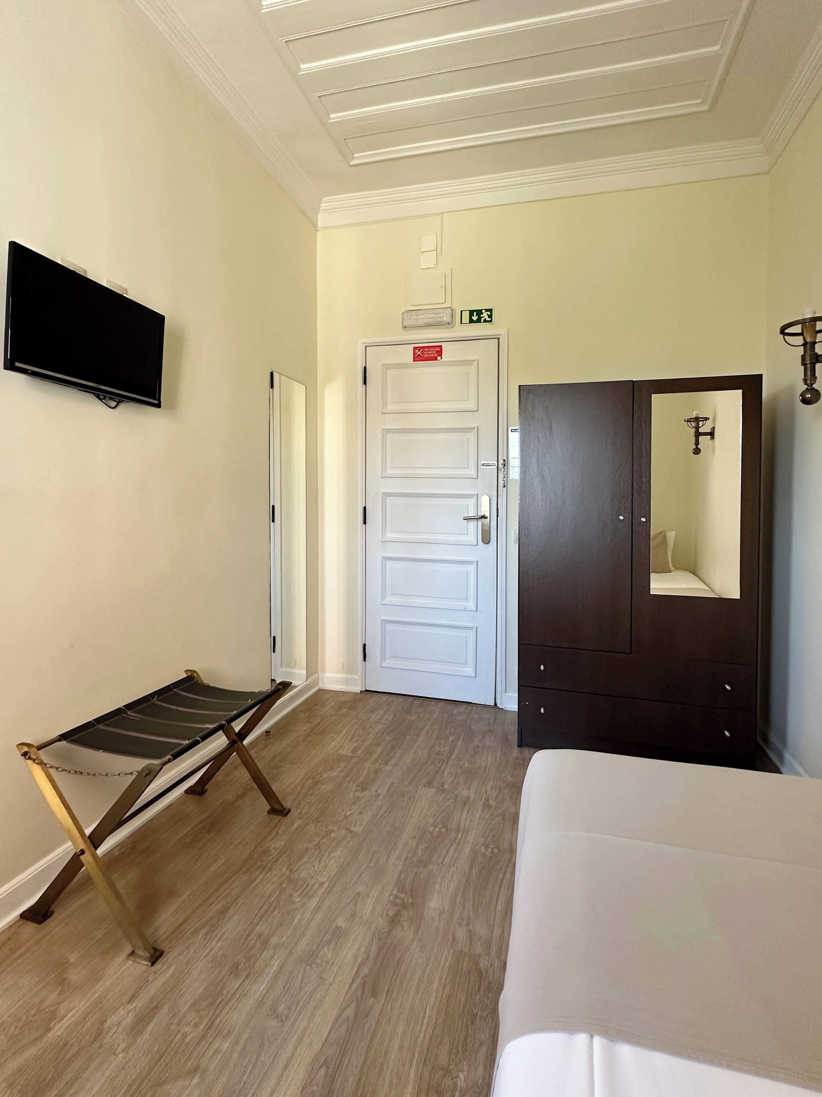 Um quarto com uma cama, uma cadeira suspensa, uma porta com sinalização de saída de emergência, uma TV na parede e um espelho.