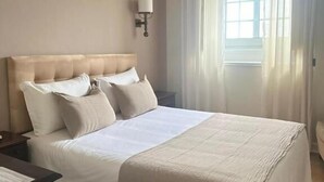 Una habitación de hotel con una cama grande, una mesita de noche, una lámpara y un ventanal con cortinas.
