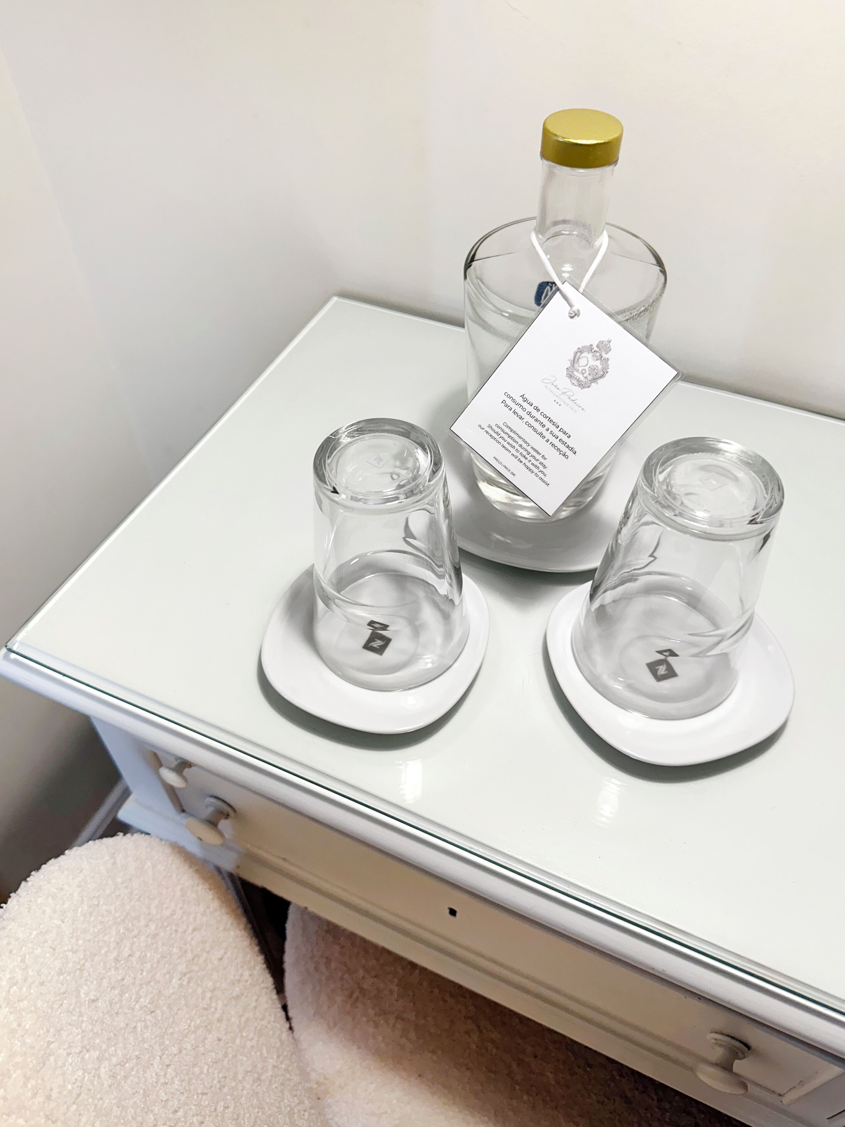 Een witte tafel met drie heldere glazen flessen en twee lege glazen bekers op schoteltjes.