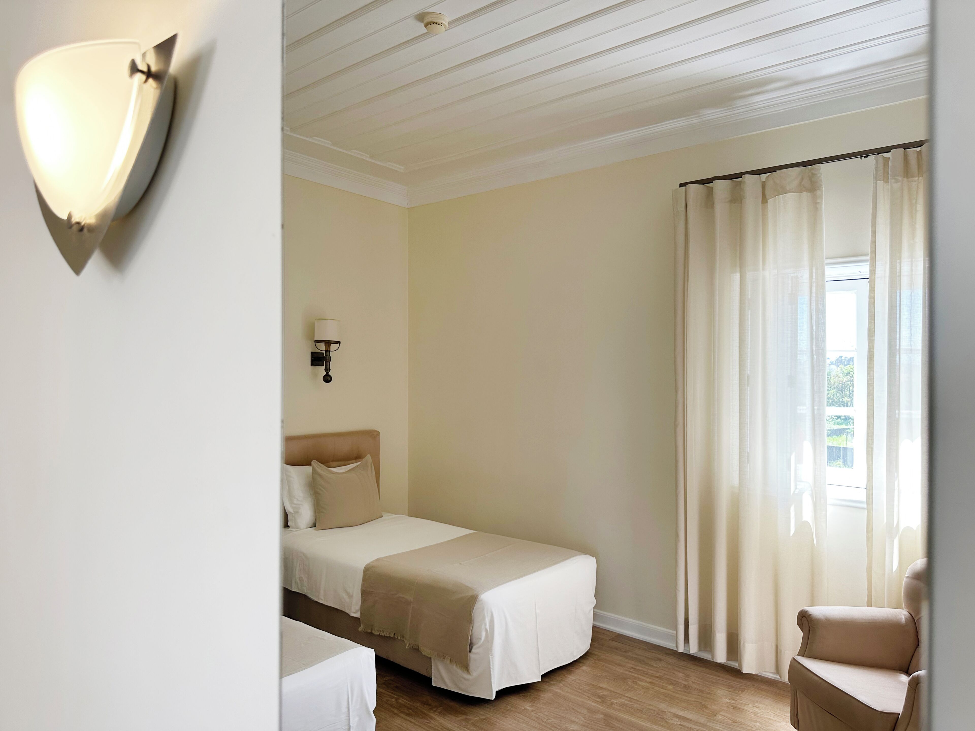 Een hotelkamer met twee bedden, een stoel, een wandlamp en een raam met gordijnen.