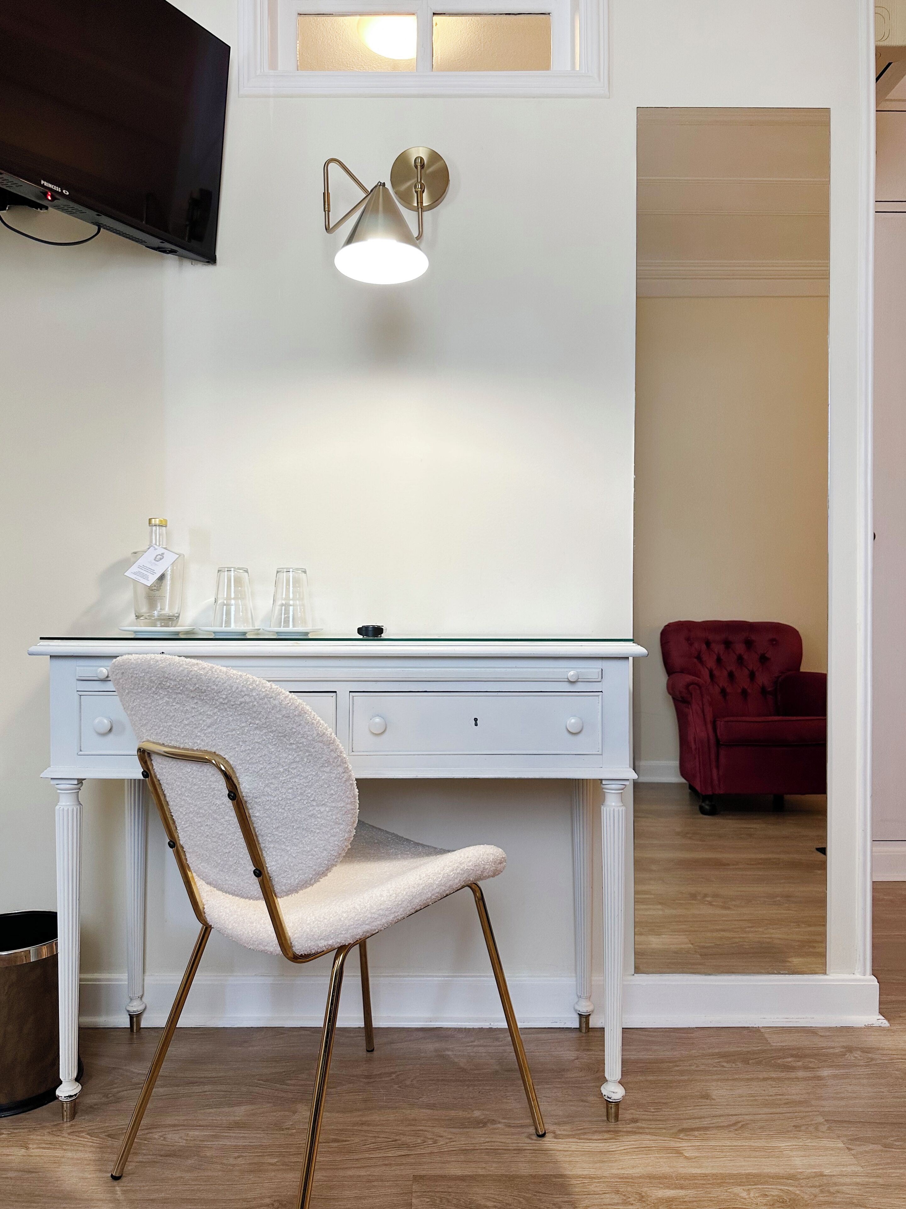Et hvidt skrivebord med glasplade, en stol med hvidt polstret sæde og gyldne ben, en vægmonteret lampe og et fjernsyn over skrivebordet.