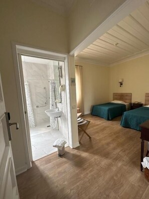 A room with a wooden floor, a bed with a green bedspread, a bedside table, a chair, a small table with a lamp, and a bathroom with a sink and shower. - Hotel João Padeiro (Cacia)