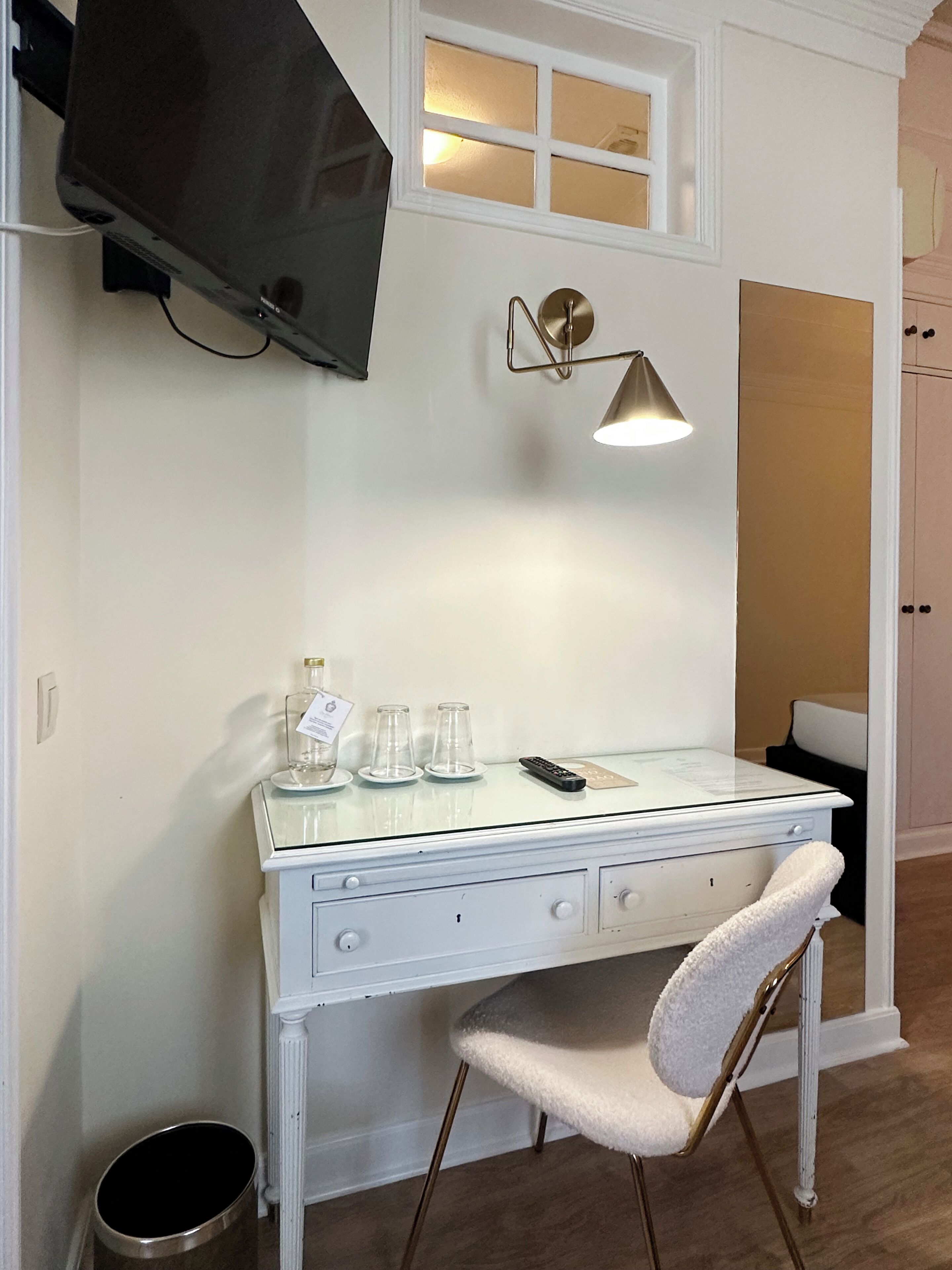 Un escritorio blanco con cubierta de vidrio, una silla, un televisor montado en la pared, una lámpara de pared y un cubo de basura pequeño.