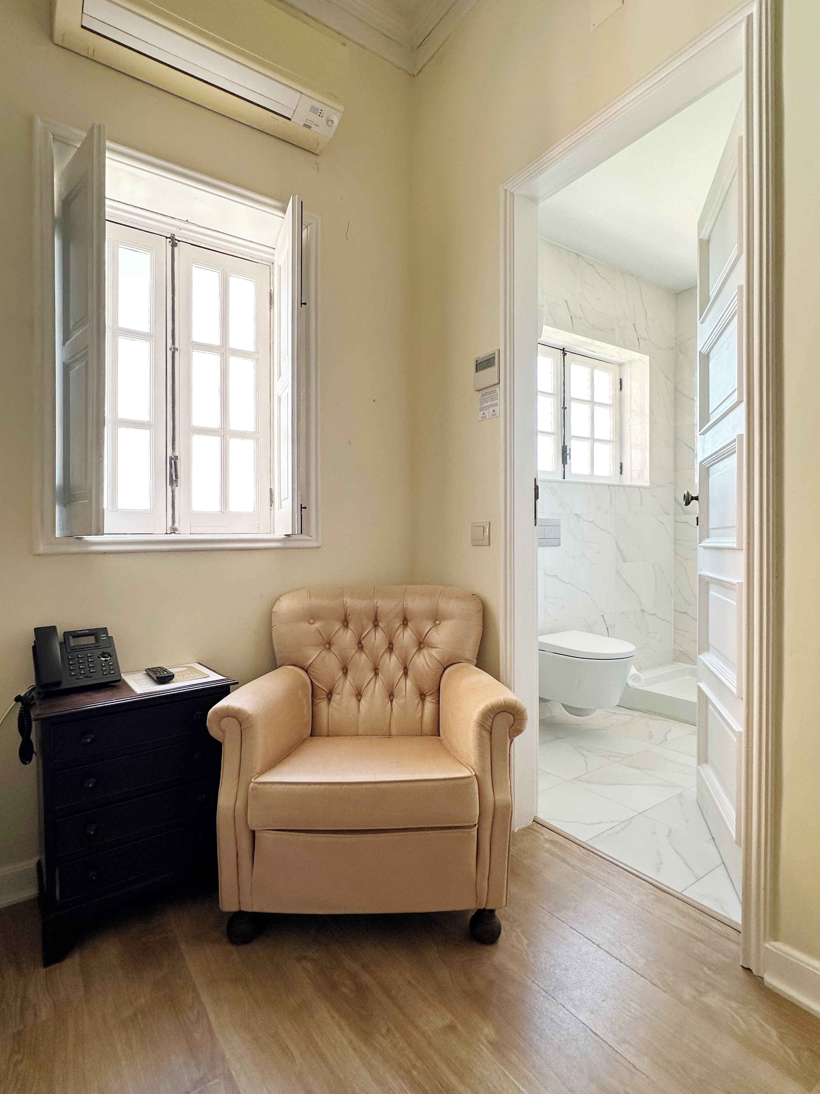 Un baño con piso de mármol, inodoro blanco, sillón color beige y mesita de noche de madera con teléfono.