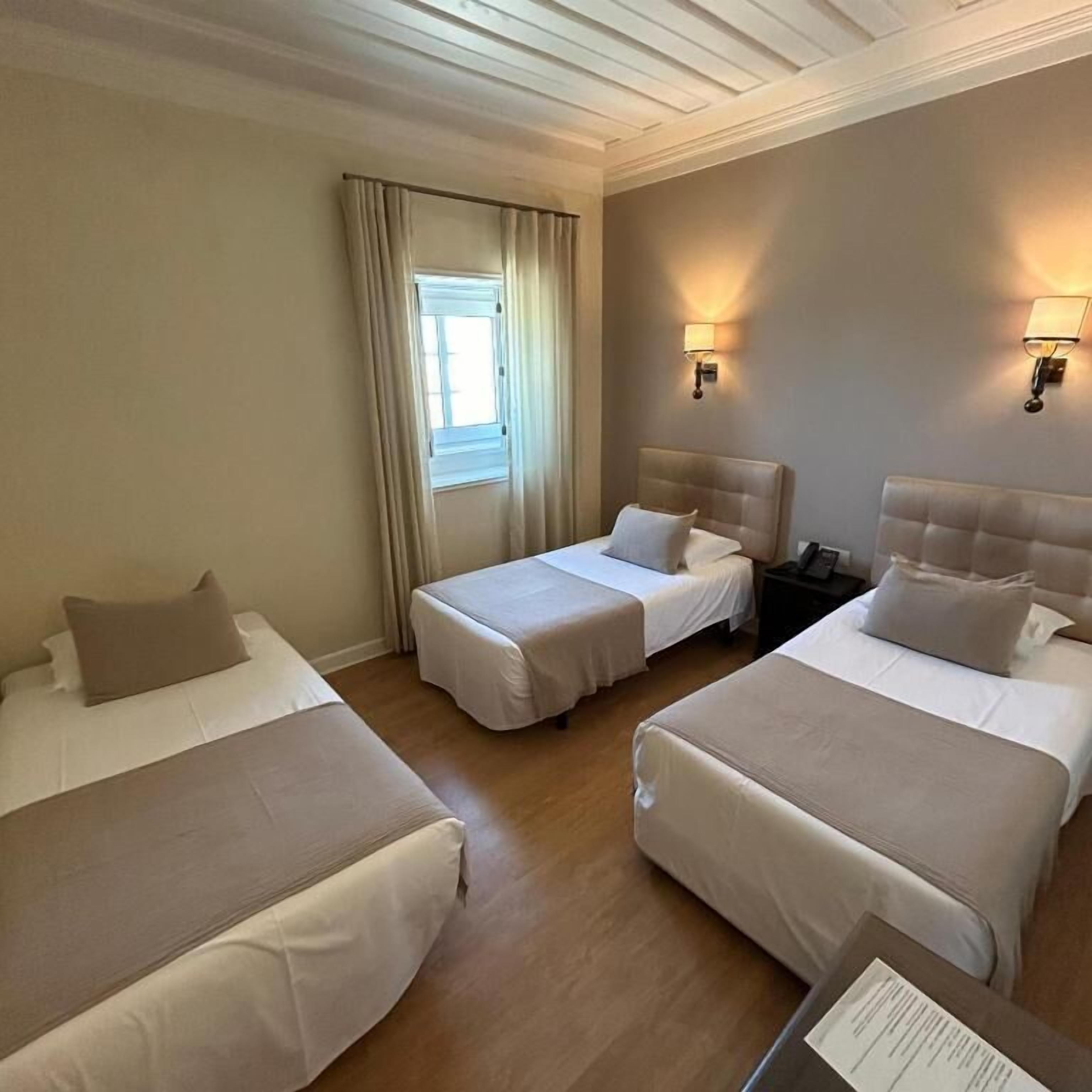 A hotel room with three beds, a window with curtains, wall-mounted lamps, and a nightstand with a lamp.