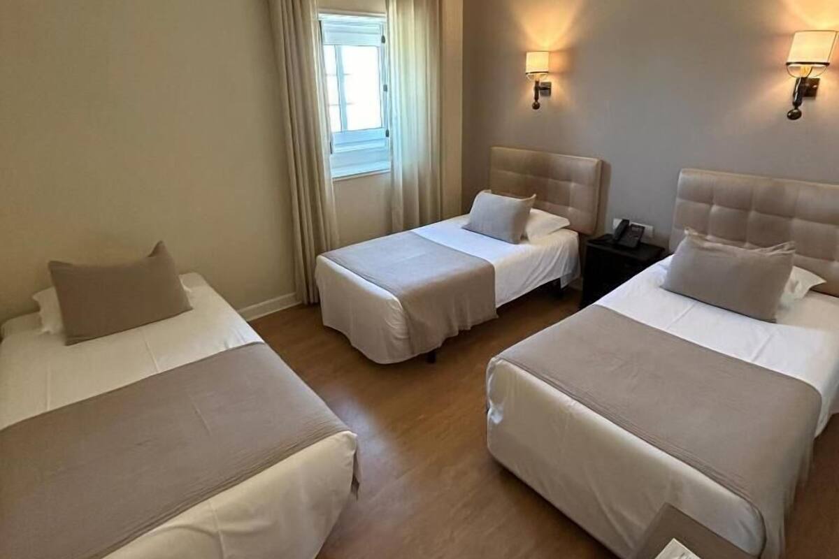 Hotellihuone, jossa on kolme sänkyä, ikkuna, jossa on verhot, seinälle asennetut lamput ja yöpöytä, jossa on lamppu.