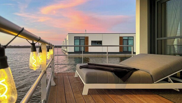 A modern outdoor lounge area with a view of a building across the water, a wooden deck, and a seating area with a cushion. - Floating house with fireplace on the lagoon (Ribnitz-Damgarten)