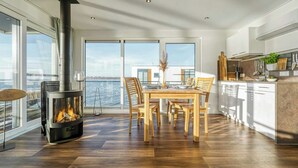 A modern kitchen with a wood-burning stove, dining area with chairs, and large windows overlooking a body of water. - Floating house with fireplace on the lagoon (Ribnitz-Damgarten)