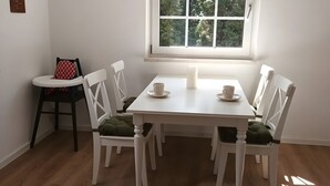 A dining area with a white table, four white chairs with green cushions, two white cups on saucers, and a window with a view of trees.