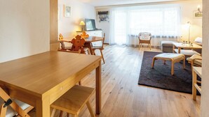Tunnelmallinen olohuone, jossa on puinen ruokapöytä, sohva, pieni televisio ja ikkuna, jossa on läpikuultavat verhot.