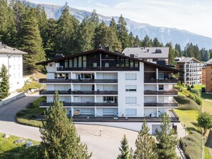 Un hotel de varios pisos con balcones, un estacionamiento y un paisaje montañoso de fondo.