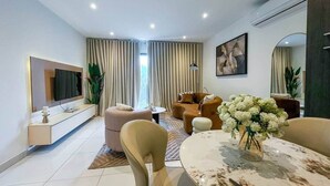 A modern living room with a sofa set, a dining table with a vase of flowers, and a flat-screen TV mounted on the wall. - Loxwood Executive 1BR | Airport City • Pool • Gym (Accra)