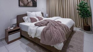 A modern bedroom with a bed, bedside table, lamp, curtains, and a potted plant. - Loxwood Executive 1BR | Airport City • Pool • Gym (Accra)