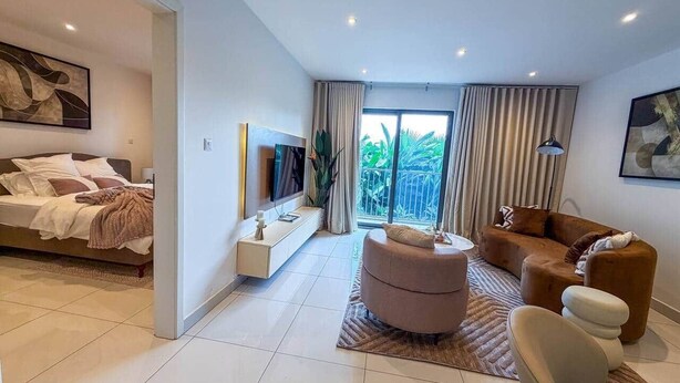 A modern hotel room with a bed, a seating area with a sofa and ottoman, a TV, a console table, and a large window with curtains. - Loxwood Executive 1BR | Airport City • Pool • Gym (Accra)