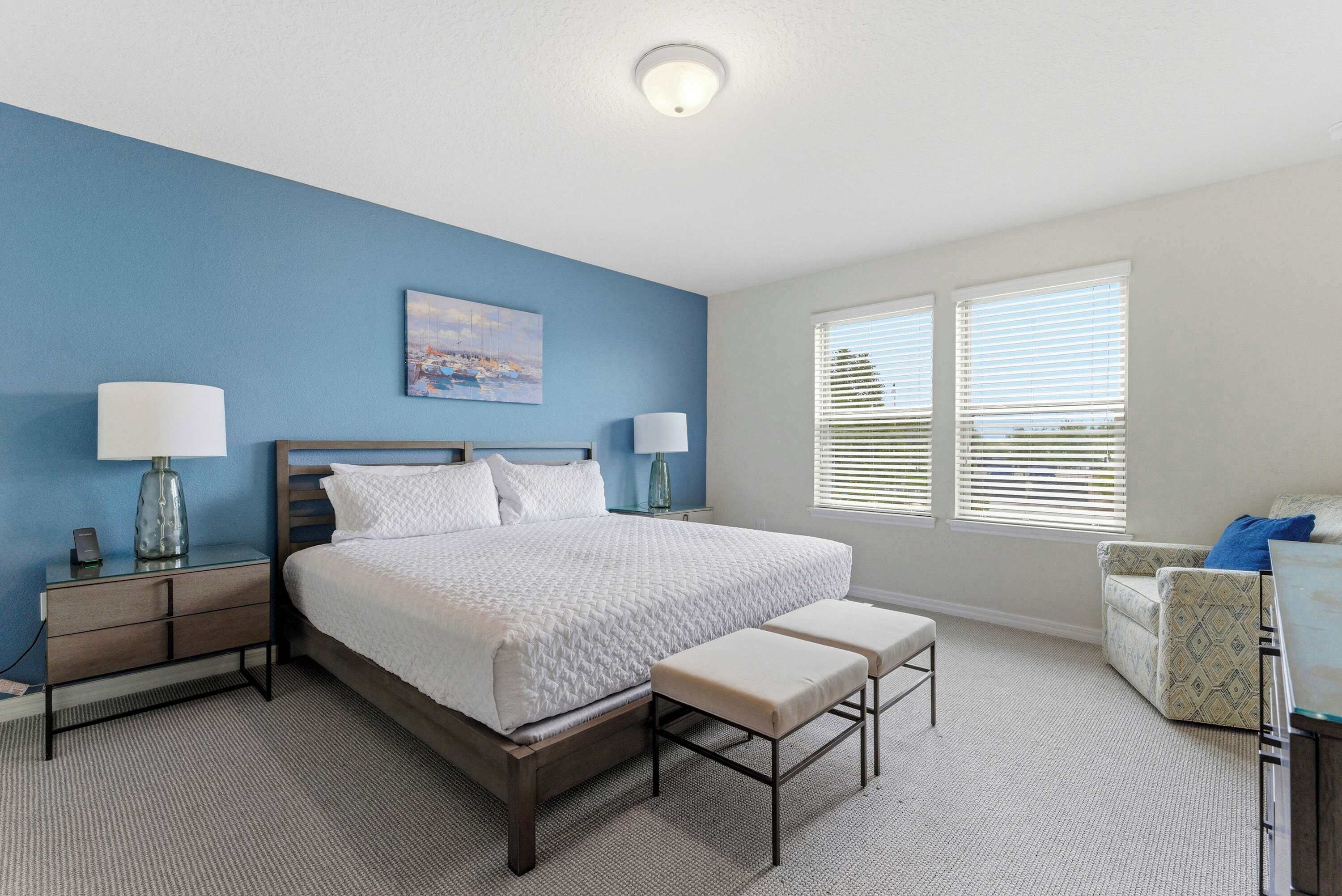 A bedroom with a bed, bedside tables, a bench, a chair, and a window with blinds.