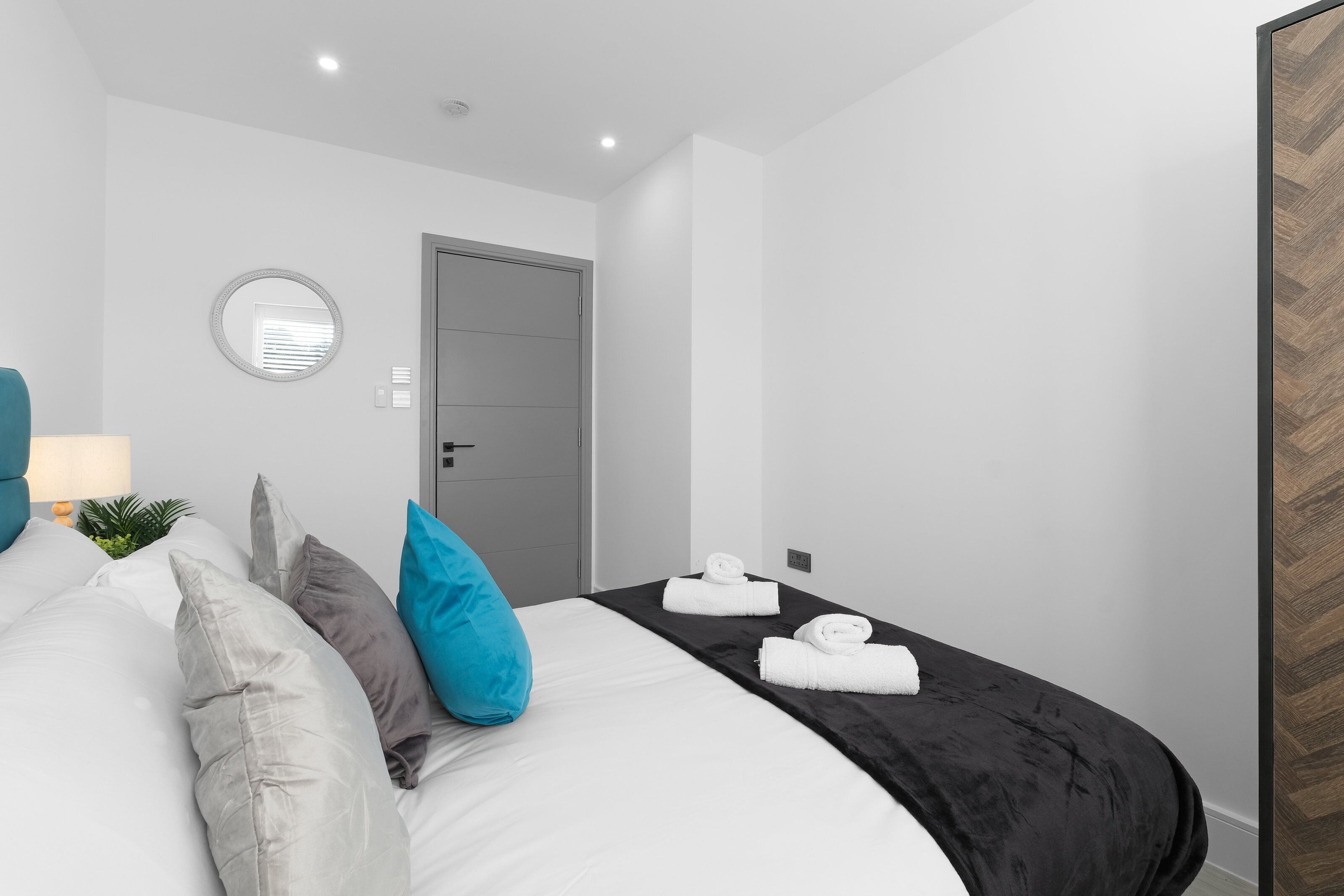 A modern hotel room with a large bed, a round mirror, a door, and a small table with a lamp.