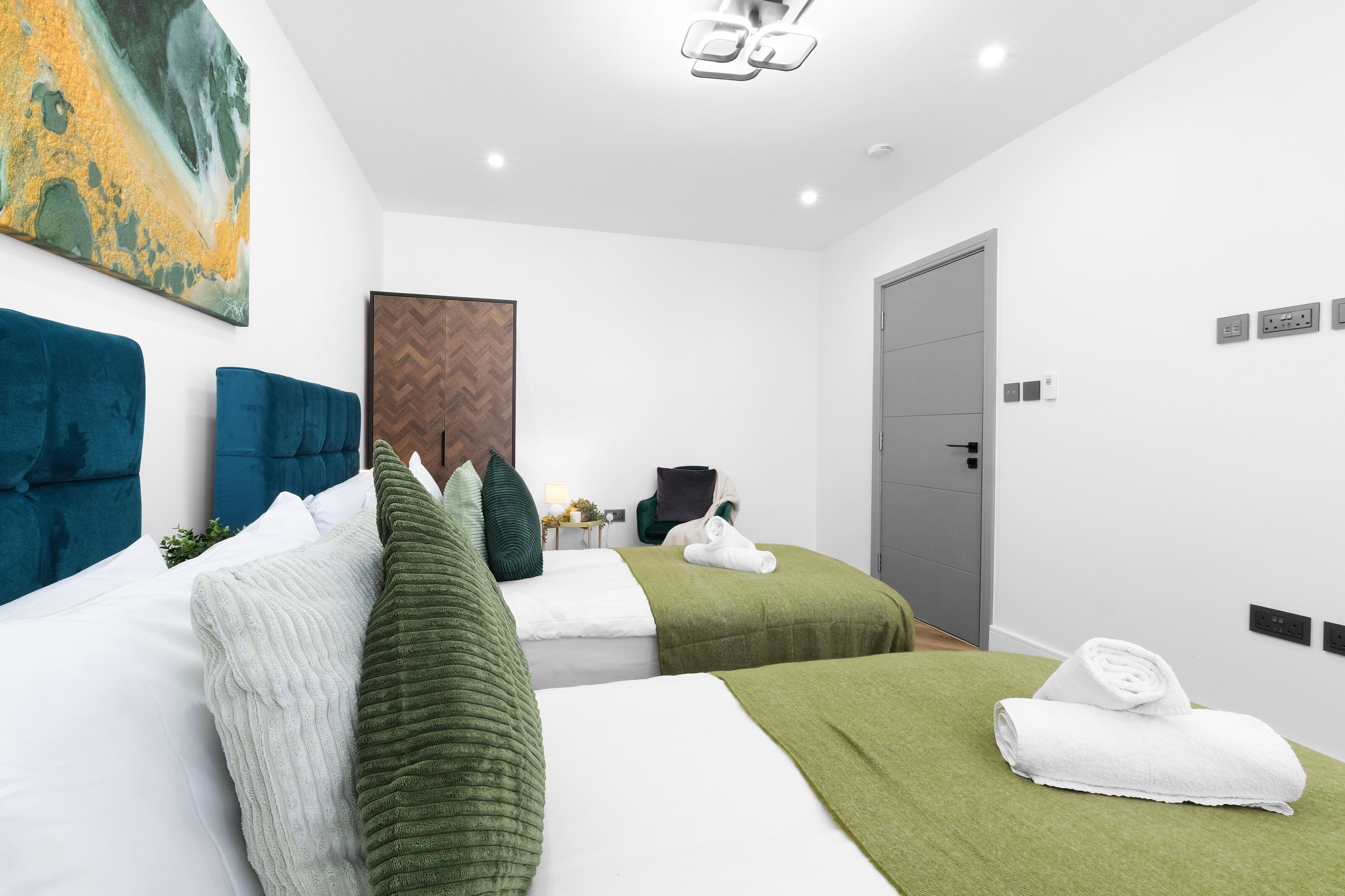 A hotel room with two beds, a large abstract painting, a wooden headboard, a door, and a wall-mounted light fixture.