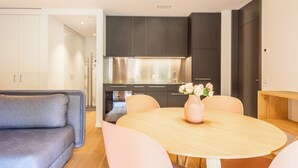 Une cuisine moderne avec des meubles de couleur sombre et des appareils en acier inoxydable, une table de salle à manger ronde en bois avec des chaises roses, et un canapé gris dans un salon lumineux.