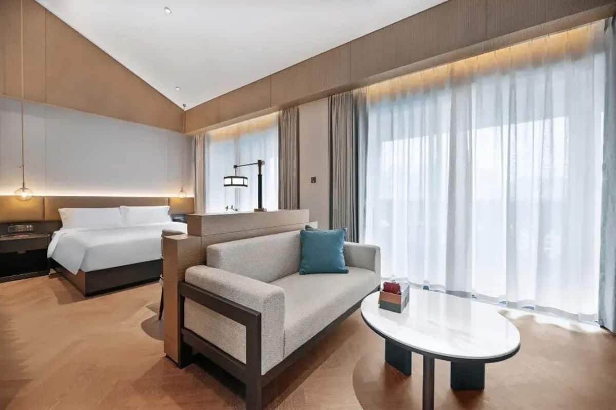 A hotel room with a bed, sofa, and round table, with large windows and curtains.