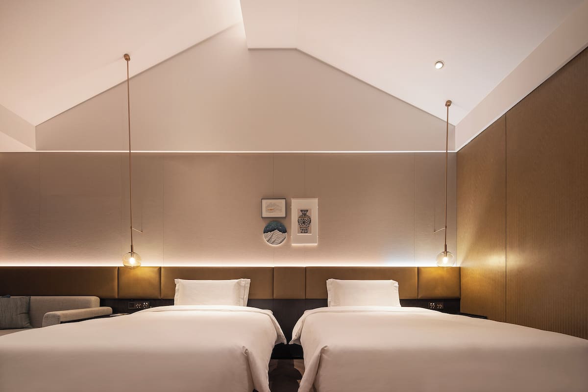 A modern hotel room with two beds, a sofa, a wall-mounted TV, and a ceiling light fixture.