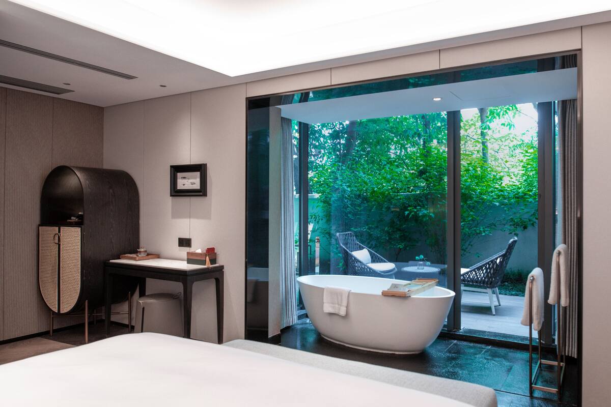 A modern hotel room with a large bed, a bathtub, a desk, and a view of a lush garden.