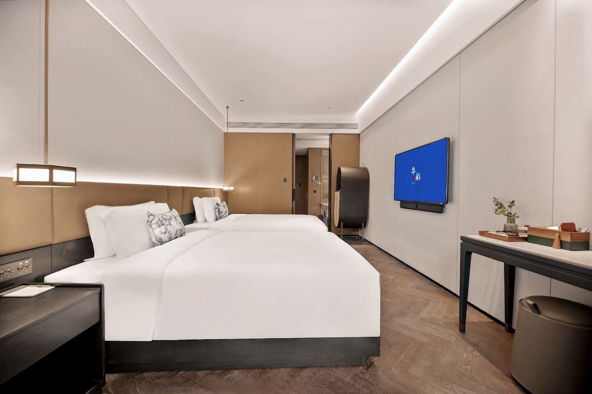 A hotel room with a large bed, bedside tables, a desk, a chair, a TV, and a trash bin.