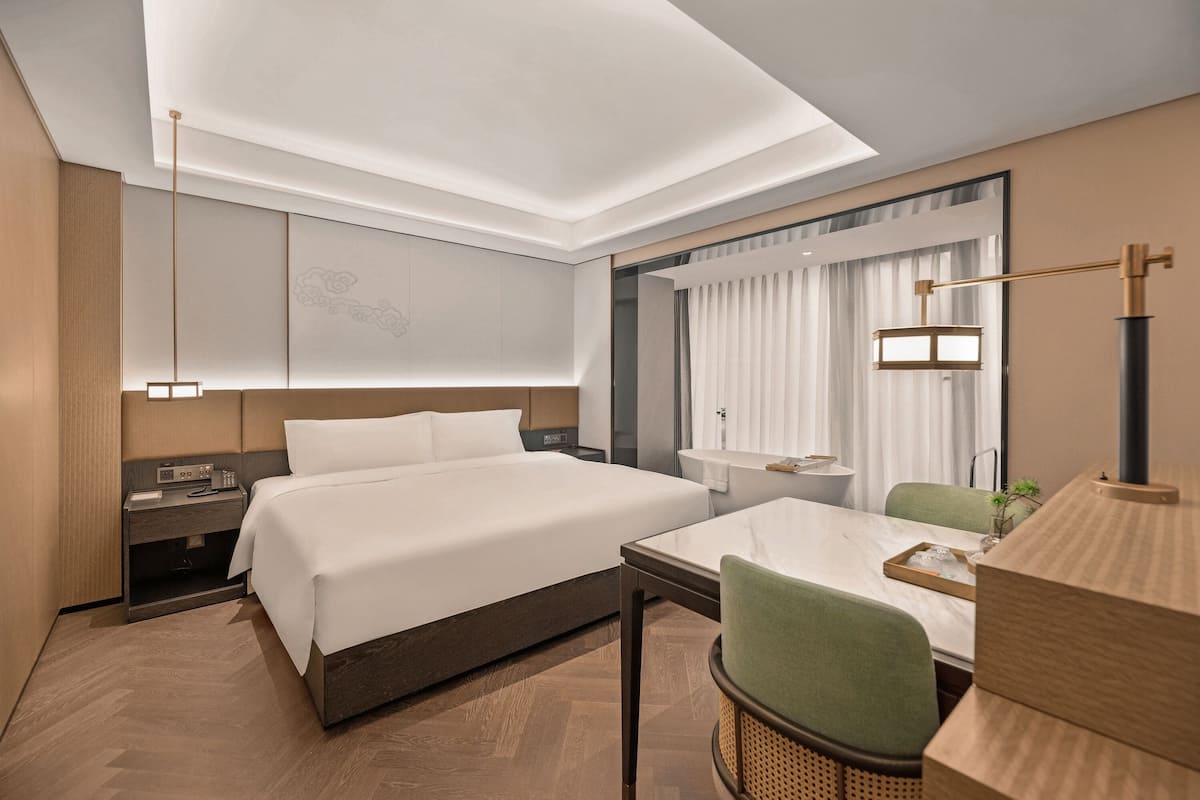 A hotel room with a bed, bedside table, lamp, chair, and a small table with a tray.