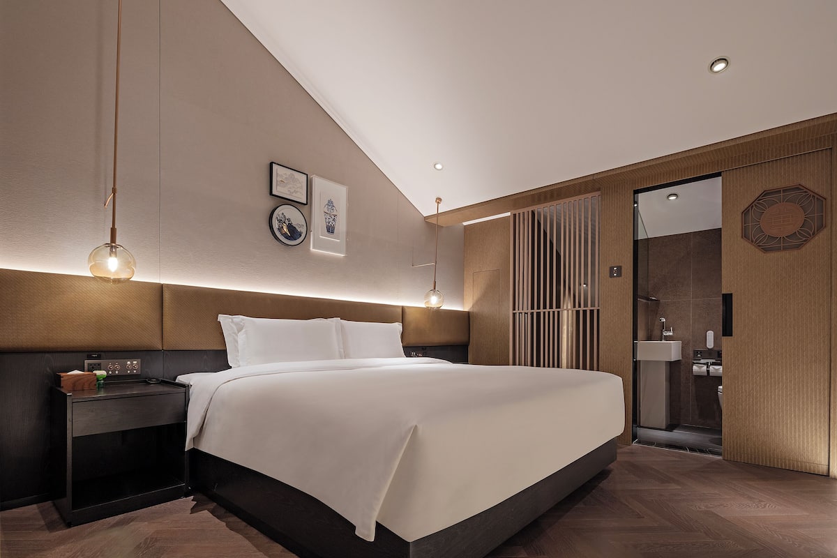 A modern hotel room with a large bed, bedside tables, a nightstand, a wall-mounted light fixture, and a bathroom visible through a glass door.