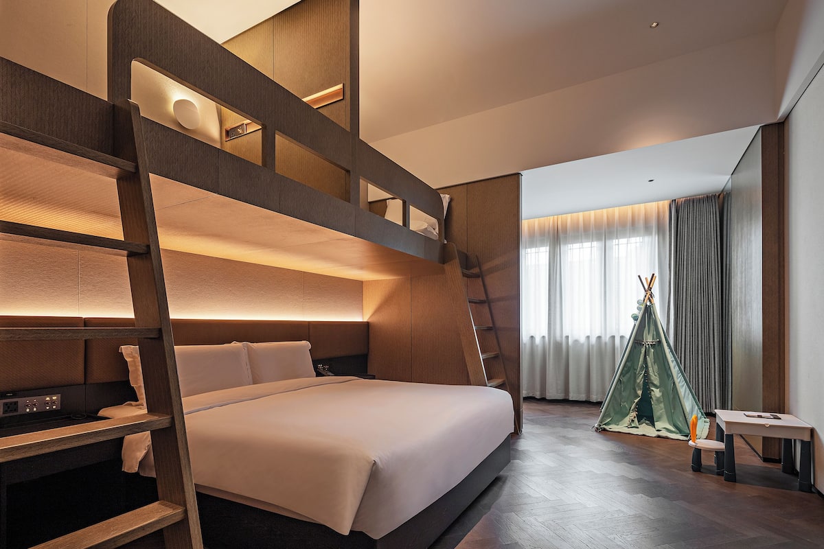 A modern hotel room with a bunk bed, a desk, a chair, a small table, and a teepee.