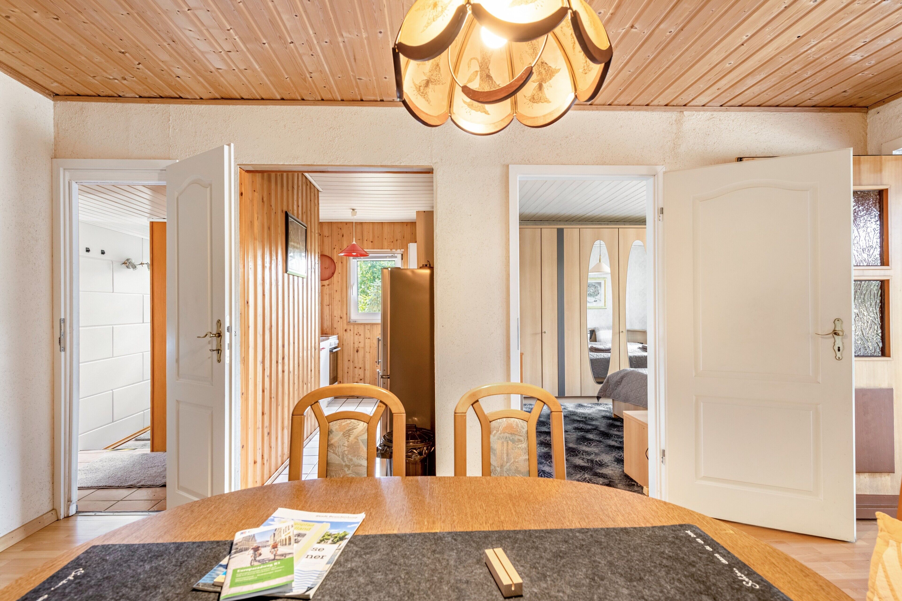 Una habitación con techo de madera, una mesa de comedor con sillas, una cocina visible a través de un umbral abierto y una cama en la habitación contigua.