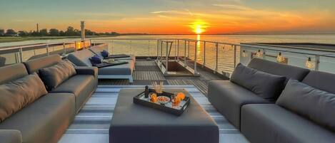 A sunset view from a deck with a sofa, a tray table with drinks, and a railing.