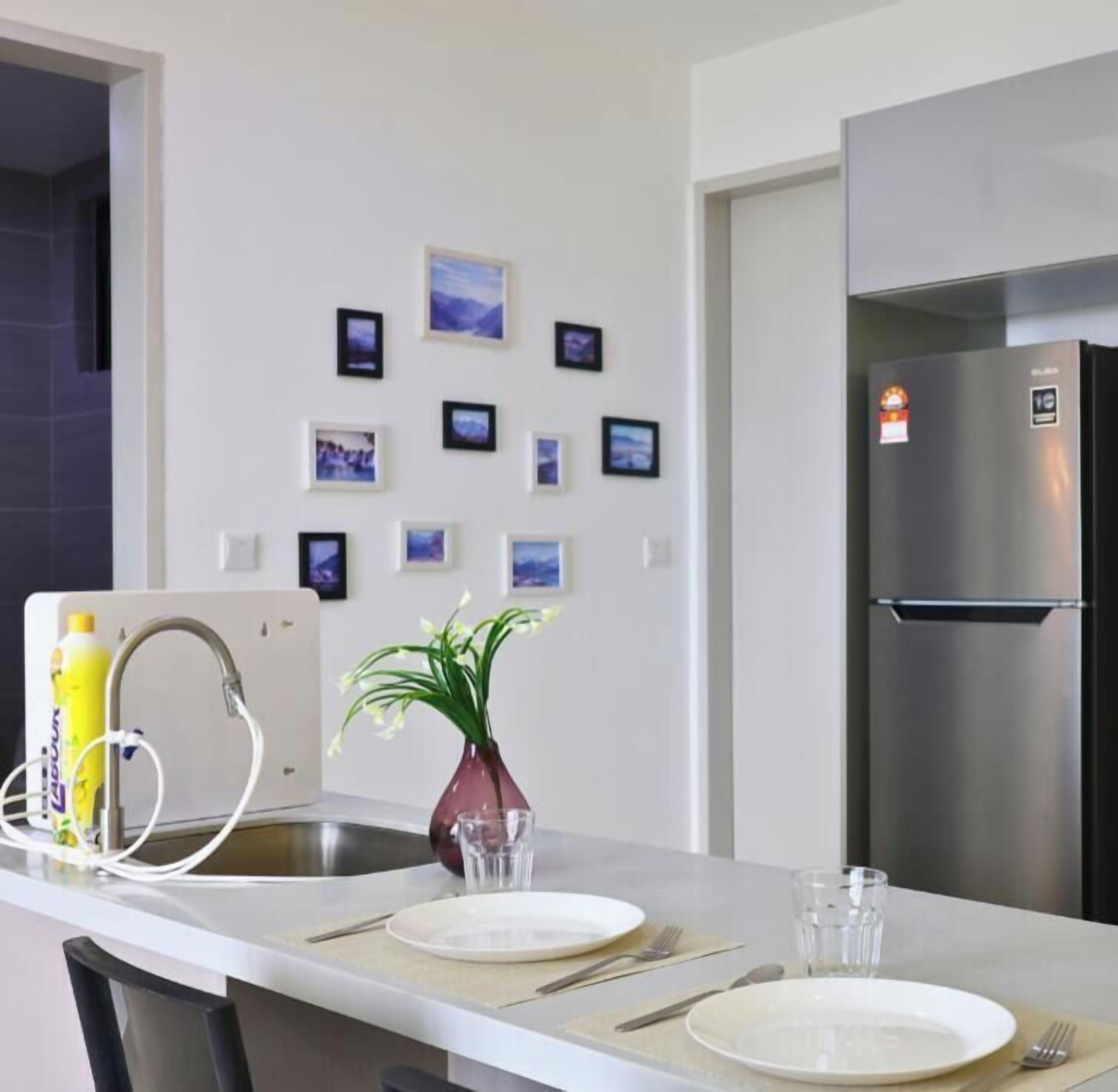 A modern kitchen with a stainless steel refrigerator, a dining table set with plates, cutlery, and glasses, and a vase with a plant on the counter.