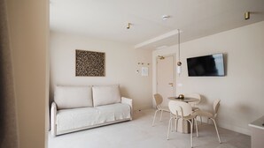 A modern living room with a sofa, a dining table with chairs, a flat-screen TV mounted on the wall, and a ceiling light fixture.