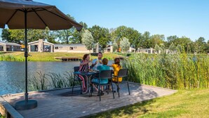 A group of people dining on a wooden deck by a lake, with a large umbrella and a view of houses in the background. - Holiday Home in Kampen With Terrace (Kampen)