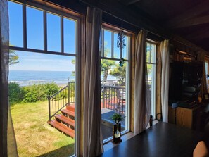 A room with large windows overlooking a beach, a staircase leading outside, and a dining area with a table and chairs. - The Pilot House at Point Bailly (Pacific Beach)