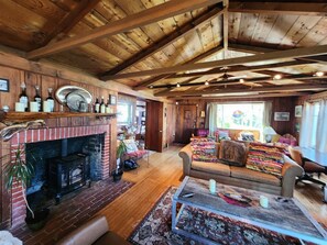 A cozy cabin interior with a fireplace, wooden beams, a sofa, and a dining area. - The Pilot House at Point Bailly (Pacific Beach)