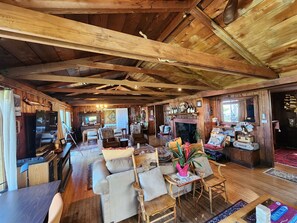 A rustic cabin interior with a wooden ceiling, a fireplace, a dining area, and a living room with sofas and chairs. - The Pilot House at Point Bailly (Pacific Beach)