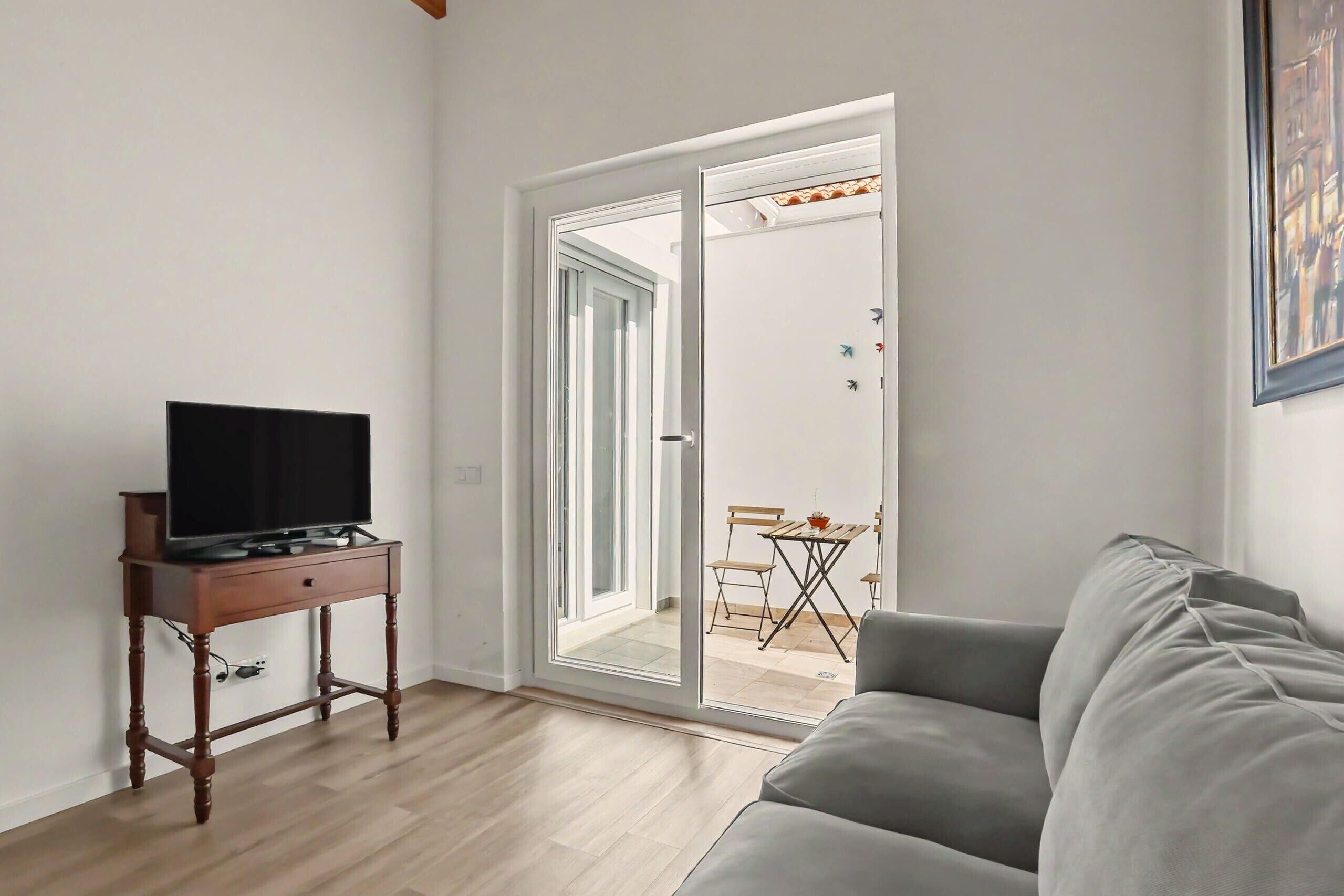En stue med et fladskærms-tv på et træbord, en grå sofa og en skydedør, der fører ud til en balkon med et bord og stole.