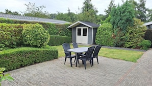 Une terrasse avec une table et des chaises, un petit abri de jardin et des haies bien entretenues.