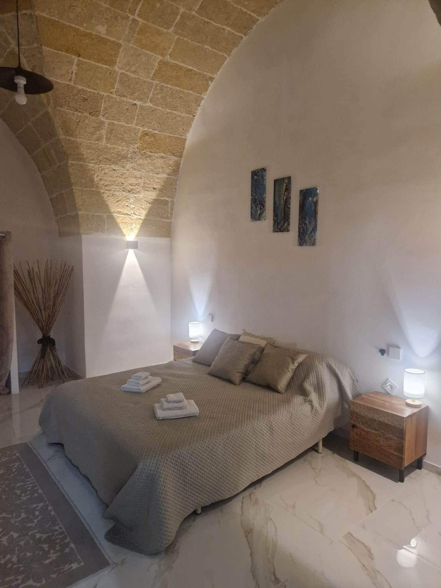 A bedroom with a large bed, a nightstand, a lamp, and a wall-mounted light fixture.