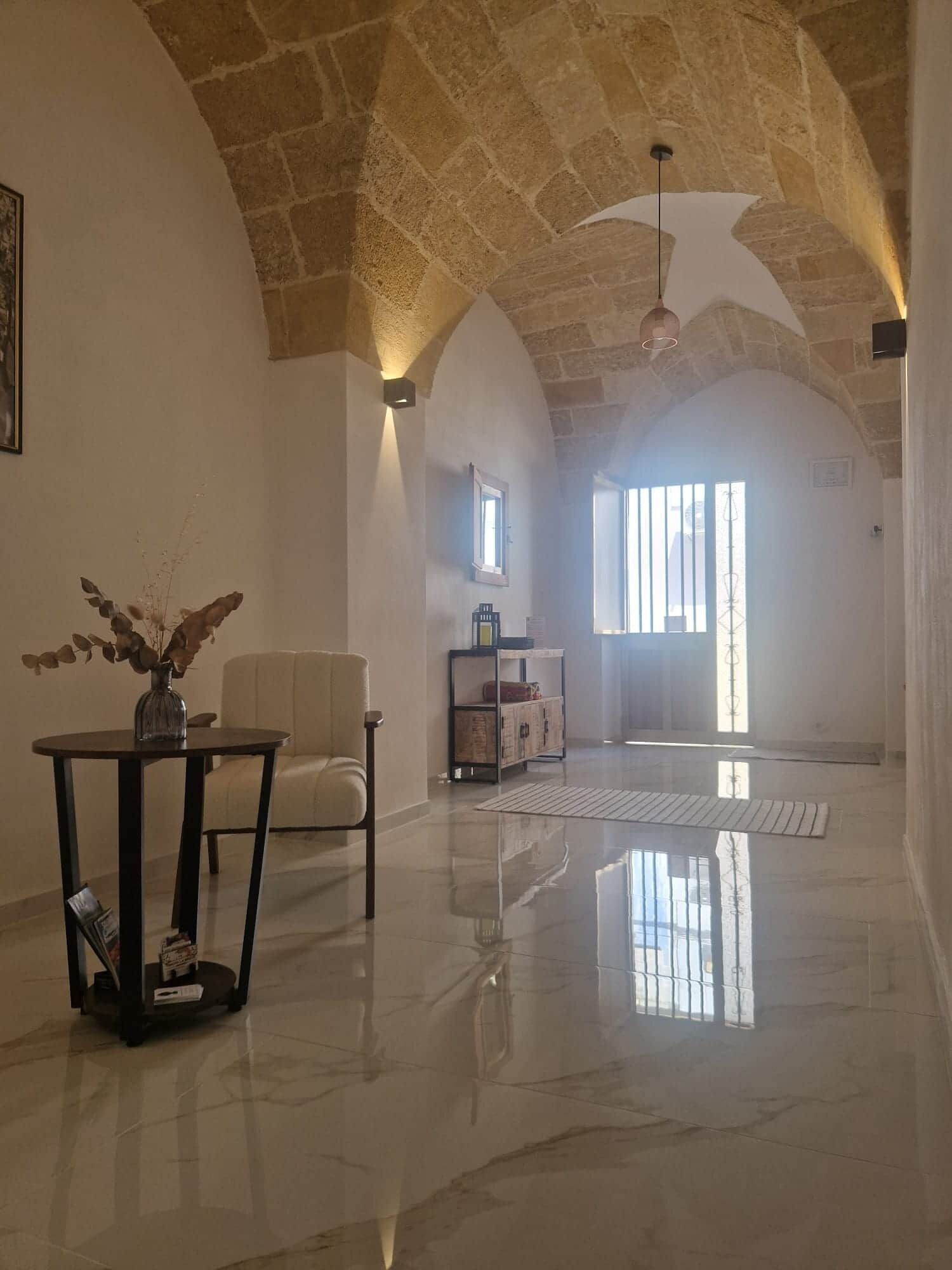 A spacious room with a high ceiling, a marble floor, a seating area with a chair and a table, and a large window allowing natural light.
