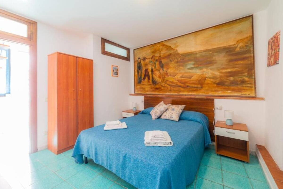 A bedroom with a bed, a wooden wardrobe, a nightstand, a painting, and a window.