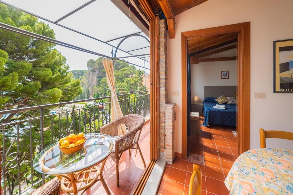 A balcony with wicker chairs, a glass table with a fruit bowl, and a view of trees and buildings.