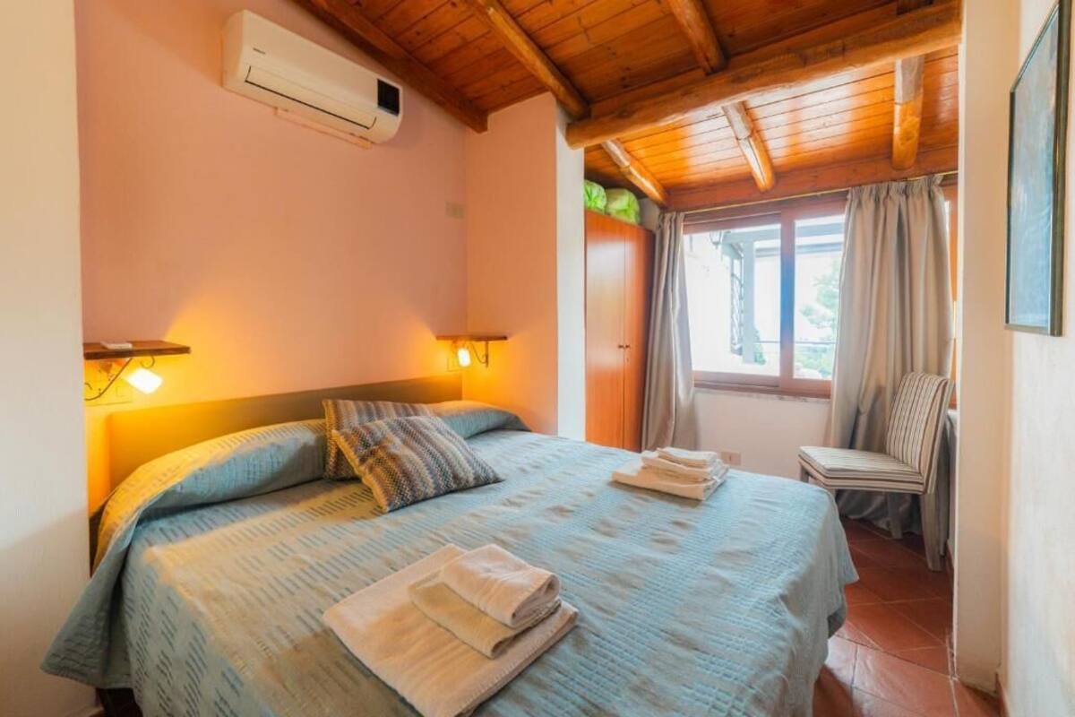 A bedroom with a bed, two bedside lamps, a window with curtains, a chair, and an air conditioning unit.