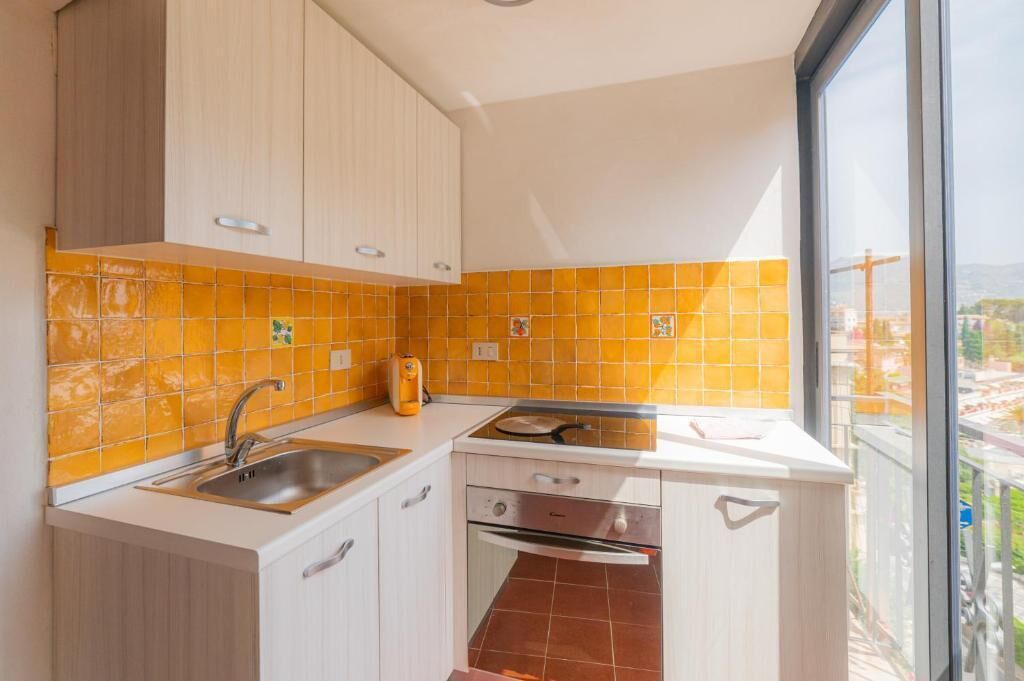 A compact kitchen with a sink, stove, and oven, featuring light wood cabinets and orange-tiled backsplash.